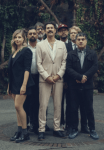 A group of seven adults, dressed in formal and semi-formal attire, pose together outdoors in front of greenery.